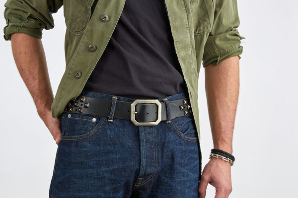 Person wearing a green jacket, black shirt, blue jeans, and a belt with pyramid studs, hole punches, and a large buckle on a white background