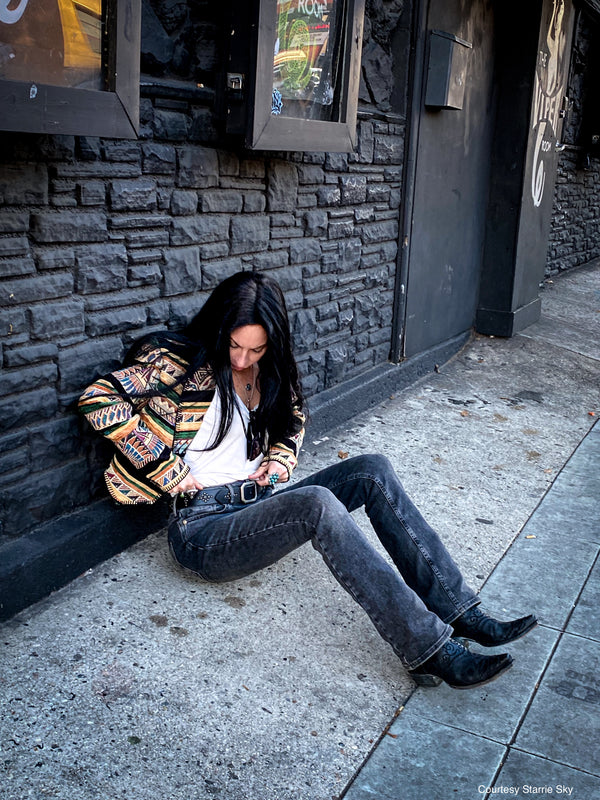 Woman sitting on a sidewalk with a colorful jacket, black jeans, and a studded black leather belt